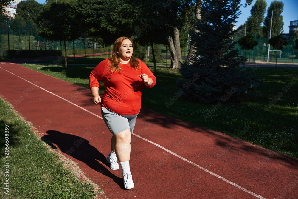 Obraz premium Curvy woman embraces fitness with joy while jogging on a sunny outdoor track