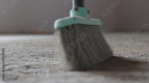 Close-up of a dusty concrete floor being swept clean with a sweeping broom in an empty indoor space. Daylight softly highlights the dust particles. Blurry background with a wall.