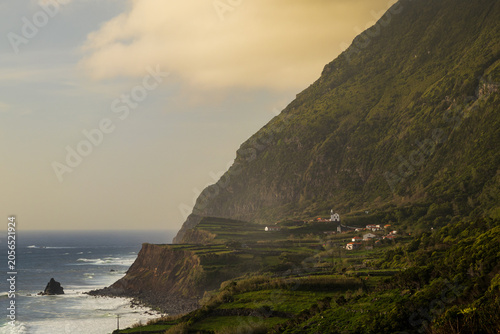 Vista panorámica de un pueblo rústico con una iglesia blanca, asentado en laderas verdes y acantilados escarpados frente al mar embravecido en la costa de la isla de Flores, Portugal.