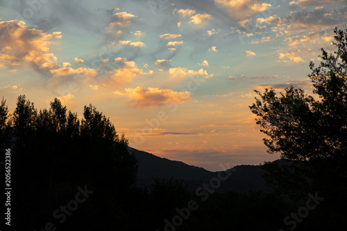 Sunset sky over dark hills and trees.Warm sunset sky with soft clouds over dark mountain hills and tree silhouettes, creating a calm natural landscape with glowing evening light and peaceful outdoor s
