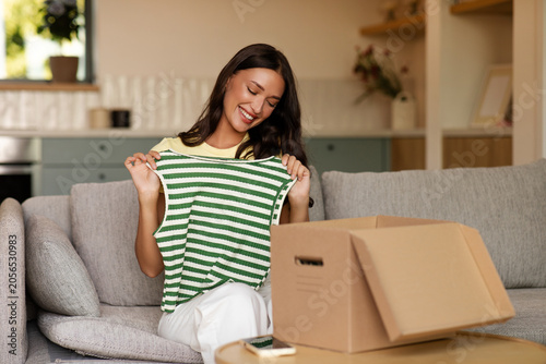 Fotografie Happy young lady unboxing cardboard parcel, trying on delivered t-shirt, taking