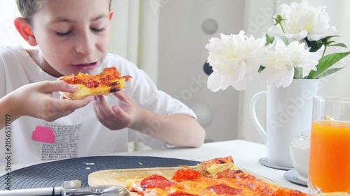 Hungry boy enjoys a slice of homemade pepperoni pizza while sitting at the kitchen table. Cozy family meal scene with fresh food, orange juice and bright natural daylight at home.