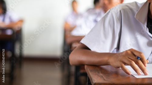 Close-up of a student's hand diligently writing on an exam paper during a school test. 
The student is using a professional pen , sitting at a wooden desk in a brightly lit classroom.