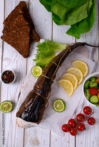 Hot smoked catfish clarias, fresh vegetable salad, rye bread, and lemon slices on parchment paper on white table. Top view. Fish delicacies