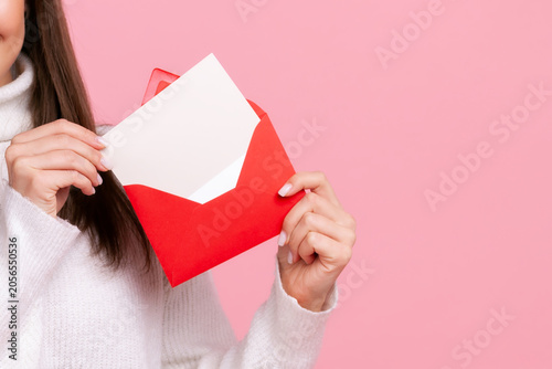 Anonymous woman holding red envelope with blank white card for invitation or romantic message isolated over pink background with copy space