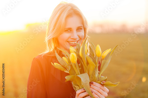 Happy blonde girl laughing and holding yellow flowers bouquet while standing in blurred field during beautiful golden hour