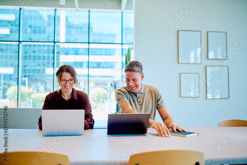 Photos Two cheerful business colleagues working on laptops, sharing a laugh and enjoyin