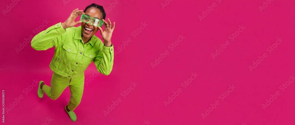 Fototapeta premium Black woman neon green outfit posing on pink background with copy space, wearing futuristic cyberpunk glasses; happy African American female style banner for modern creative fashion marketing project.