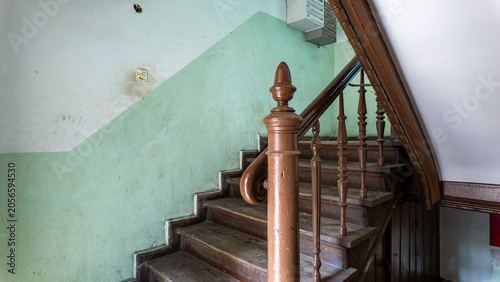 Fotografie Interior of an old tenement house staircase featuring worn wooden stairs, a brown railing with turned balusters, and a wall painted with a green dado panel under natural daylight