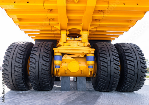 Low-angle view of the underside of a large yellow dump truck focusing on dual rear axles, heavy tread tires, suspension components and metal chassis. Bright