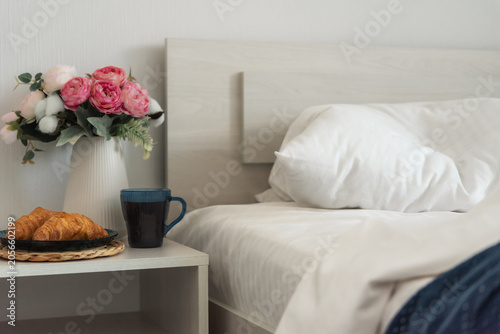 Croissants and coffee on a bedside table next to an unmade bed in a bright bedroom interior. Cozy morning, hospitality and accommodation concept.
