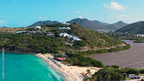 Aerial view of Friar's Bay and Etang Guichard salt pond on the French side of Saint Martin island in the Caribbean. White sand beach organized with beach umbrellas, sunbeds, and people relaxing.