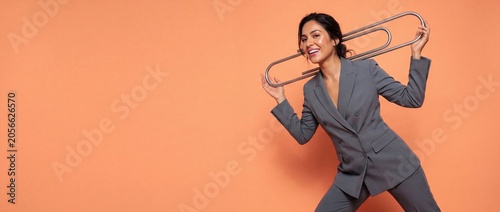 Businesswoman holding giant paperclip smiling on orange background as creative office organization concept with copy space; professional female executive in gray pantsuit carrying big paper clip.