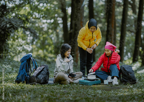 Group of diverse friends hikers asian female wear puffer jacket goose down in a forest. They are cooking food and drinks with portable stove in camping ground surrounded by trees and greenery.