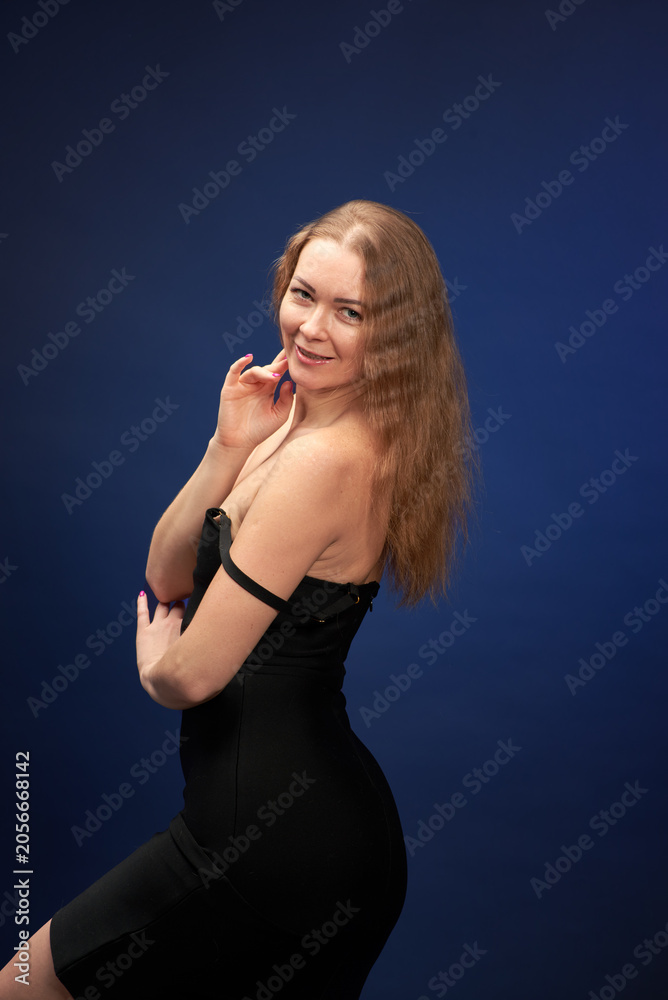 Fototapeta premium A woman with a beautiful figure in an elegant black dress poses against a dark blue background.