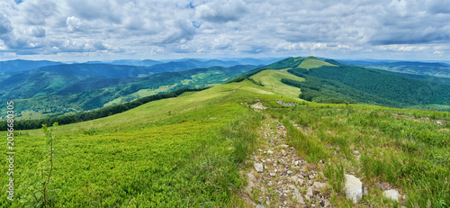 A dirt trail leads across a lush green mountain ridge toward a grassy peak, all under a vast, dramatic blue sky filled with bright, shifting white and grey cumulus clouds.
