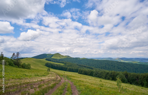 A dirt trail leads across a lush green mountain ridge toward a grassy peak, all under a vast, dramatic blue sky filled with bright, shifting white and grey cumulus clouds.