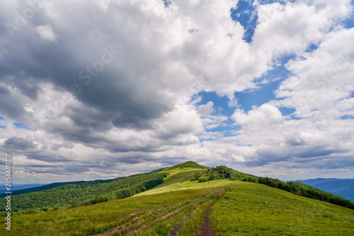 A dirt trail leads across a lush green mountain ridge toward a grassy peak, all under a vast, dramatic blue sky filled with bright, shifting white and grey cumulus clouds.