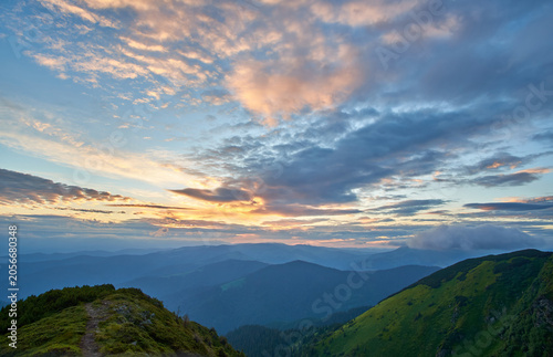 A breathtaking sunset glows over rolling mountain silhouettes, with foreground spruce trees and lush greenery standing under a dramatic, cloud-filled twilight sky in this serene alpine landscape.