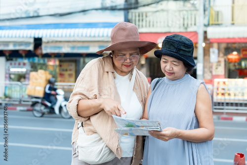 Senior asian women travelers standing on street checking map and smartphone planning route enjoying retirement lifestyle vacation together exploring culture friendship during joyful urban journey