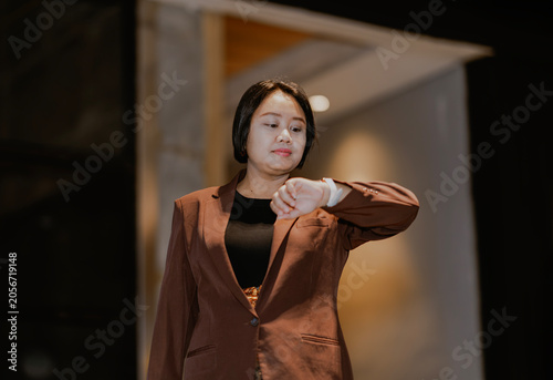 Adult Asian businesswoman wearing a brown suit looking her wristwatch standing in a luxury office building indoors.