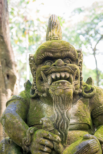 Konstfotografi A medium close-up vertical shot captures a mossy, hand-carved Balinese stone statue of a fierce, fanged guardian spirit sitting in a tropical forest setting in Ubud, Bali