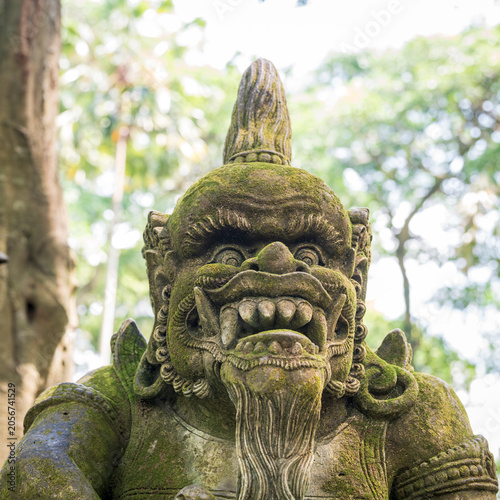 Foto A close-up vertical shot features a moss-covered stone statue of a fierce, fanged Balinese guardian deity standing against a soft-focus forest background in Ubud, Bali