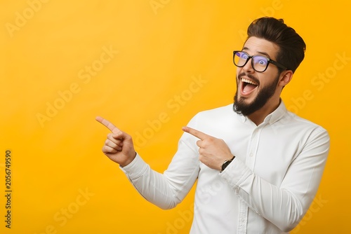 Excited young man pointing aside at copy space, happy businessman showing product on yellow background, cheerful male professional in white shirt