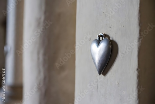 a small silver ex-voto heart hanging alone on a plain chapel wall, symbolizing gratitude, healing, and silent answered prayer