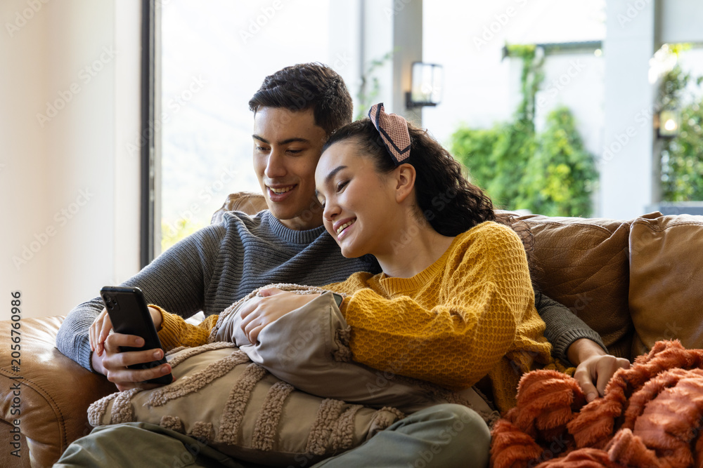Obraz premium Diverse couple sitting together in living room, leaning and looking at smartphone on leather sofa