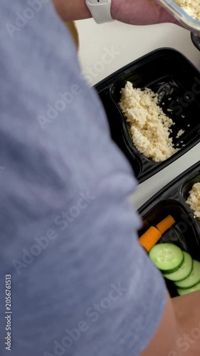 Vertical video: Lifting bowl, cook in blue tee scooping grain into black trays on counter for meals