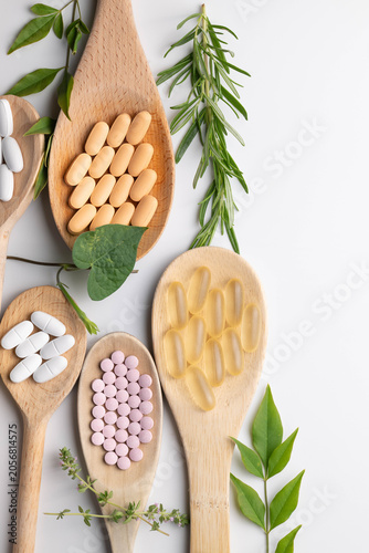 Wooden spoons holding various health supplements, soft gels, capsules, tablets, on white background.  Fresh green leaves, vines, and herb accents. vertical.