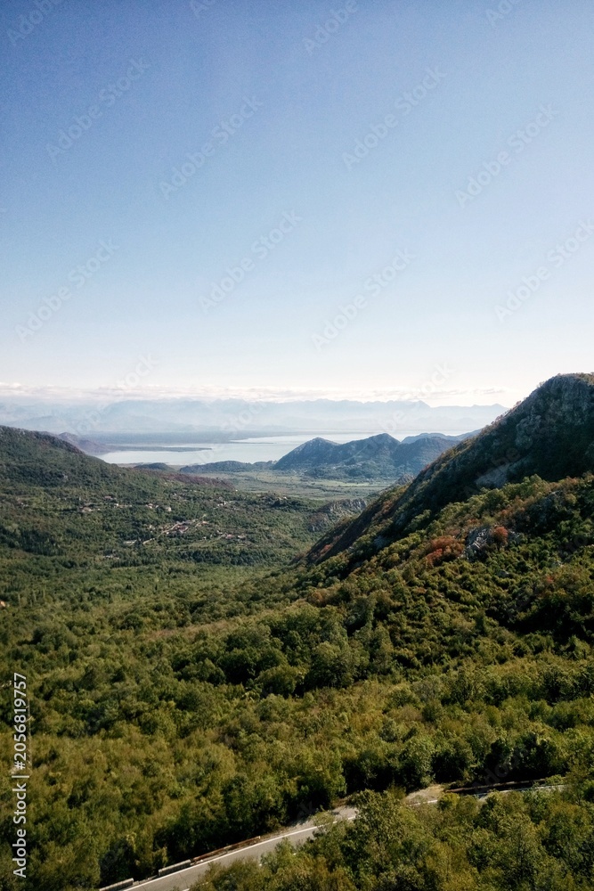 Fototapeta premium High angle panoramic view of Skadar lake valley in Montenegro with green forest trees on mountain hillsides and asphalt road asphalt curve line path in foreground and clear blue sky horizon background