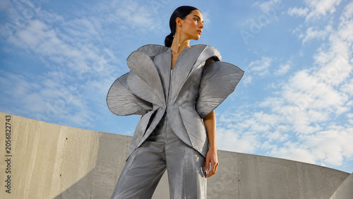 Futuristic woman in a sculptural silver couture outfit posing against modern concrete architecture and a cloudy blue sky.