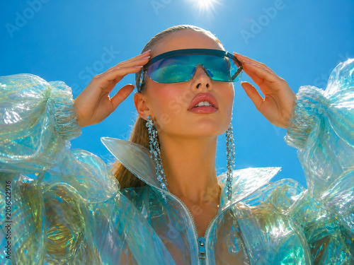 Futuristic fashion portrait of a woman in an iridescent translucent jacket and oversized visor sunglasses under a bright blue sky.