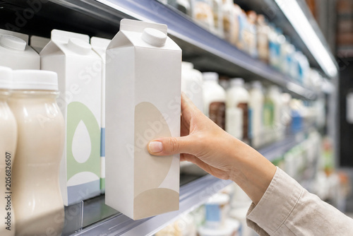 Close-up of a shopper selecting a blank milk carton from a refrigerated dairy shelf in a supermarket aisle.