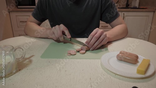 Person chopping sausage on green cutting board, focused hands slicing rounds beside plate with whole sausage and cheese, kitchen counter under warm light, casual home cook preparing simple snack