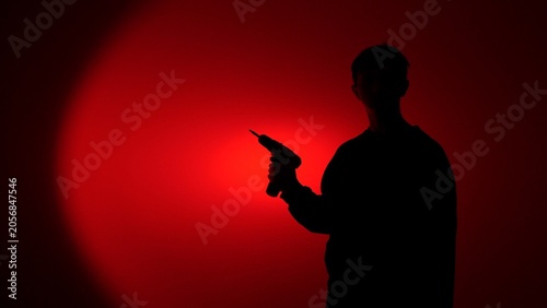 Bilde på lerret Dark male silhouette grips a power drill, backlit by an intense red spotlight, e