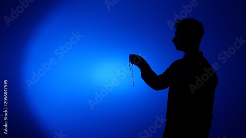 фотографія Silhouette of man holding a rosary and cross against a blue background, evoking