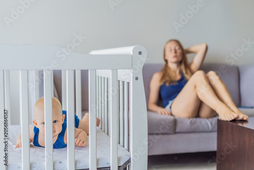 Fotografia Very tired mother sitting by baby crib at home feeling exhausted