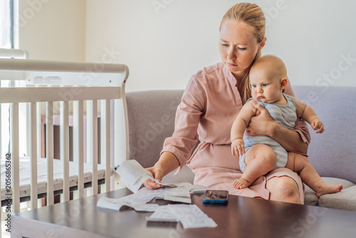 Mother sitting on sofa holding baby while calculating monthly expenses with receipts at home. Stressed mom managing budget and childcare. Financial stress, parenthood and real life concept