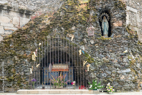 Grotto of the Immaculate Conception in Viveiro, Galicia, Spain. Stone shrine with Our Lady of Lourdes statue, arched iron gate, ex-voto offerings, and fresh flowers in a mossy rock wall.