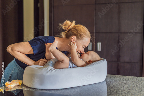 Mother talking to baby lying in cozy lounger on kitchen table while cooking at home. Caring mom interacting with infant during daily routine. Parenting, multitasking and family life concept