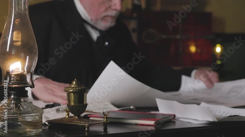 1800s man in his office unrolls a large drawing with focus on oil lamp