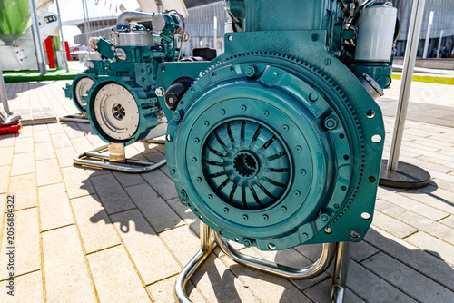 A large weathered cast-iron industrial pump painted turquoise with peeling paint sits on a paved plaza under bright sunlight. The circular pump housing