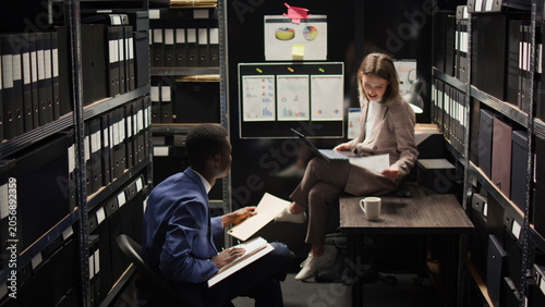 фотографія In room crammed with clues and records, Caucasian female investigator sits on table analyzing evidence with police officer, African American inspector hands over case data to policewoman with laptop