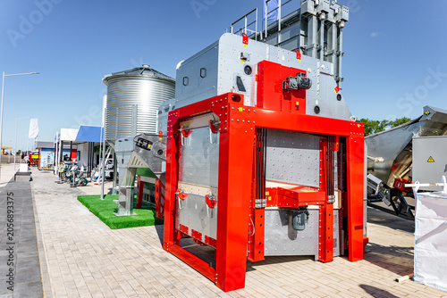 Fotografía A bright red industrial machine displayed outdoors on a paved exhibition area, surrounded by other metallic equipment and kiosks