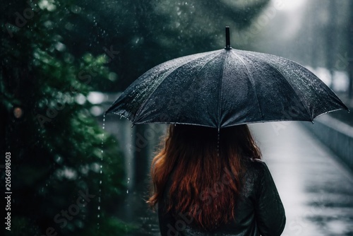 A woman with long red hair stands under a black umbrella during a rainstorm. The scene captures a melancholic and moody atmosphere in a park. Perfect for emotional or weather related content.