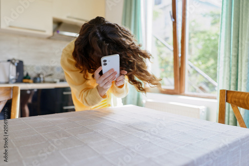 Fotografía Young woman with wavy hair in a yellow sweater using a smartphone at a kitchen table