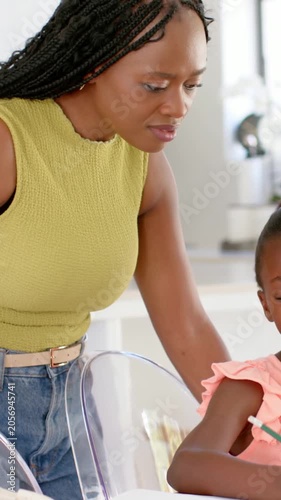 Vertical video: Fixing clear chair, mom in green leaning over girl at table guiding with notebook